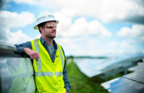 A solar installer wearing a hardhat, sunglasses and a high-visual vest leans on an installed row of solar panels.