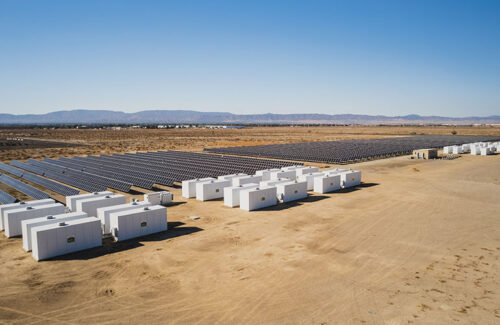 White energy storage units containing electric vehicle batteries sit parallel to a field of solar panels.