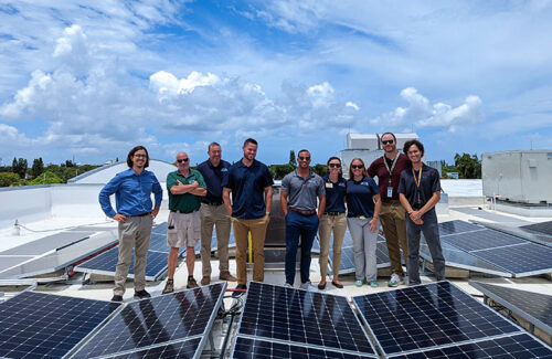 A group of nine people stand shoulder to shoulder between panel rows of a rooftop solar project. 