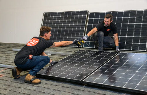 Two people crouch on a section of rooftop used for training modules and demonstrate how to install solar panels. 